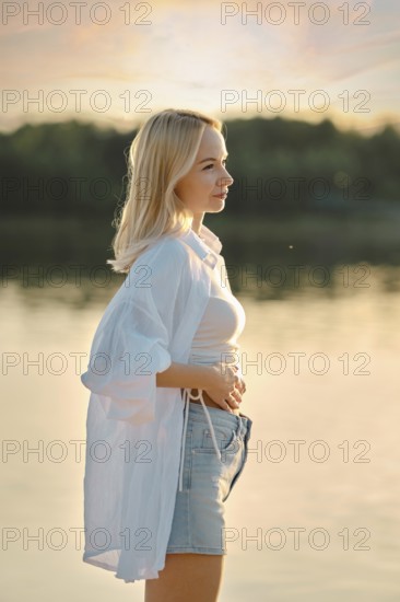 A young woman stands by a calm lake during sunset, feeling the gentle breeze. Her casual outfit reflects the relaxed atmosphere, with the sun casting warm hues on the water and surroundings