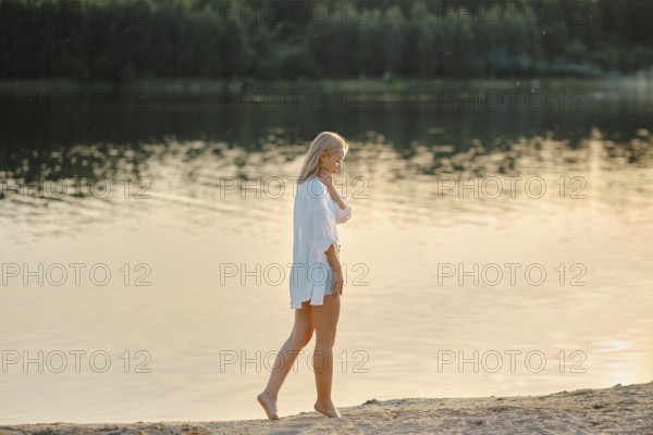 Pensive woman strolls barefoot on the sandy beach beside a serene lake. Golden sunlight reflects off the water as trees stand in the background, creating a calm and soothing atmosphere during sunset
