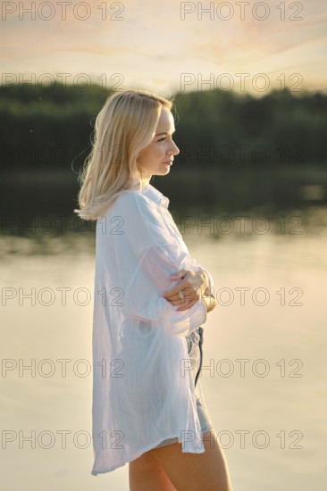 A young woman stands pensively by a calm lake at sunset, dressed in a light shirt and shorts. The warm glow of the setting sun reflects on the water, creating a peaceful atmosphere