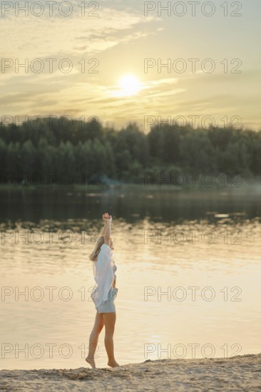 A woman stands on the sandy shore of a tranquil lake, raising her hand joyfully towards the setting sun. The golden light reflects off the water, surrounded by lush greenery
