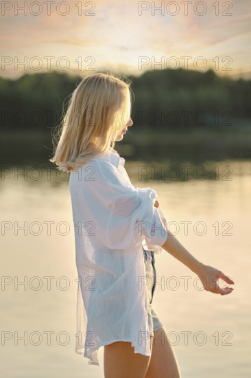 A young woman stands by the tranquil lake at sunset, wearing a white shirt and denim shorts. The soft sunlight casts a warm glow, highlighting her relaxed demeanor against the serene water backdrop