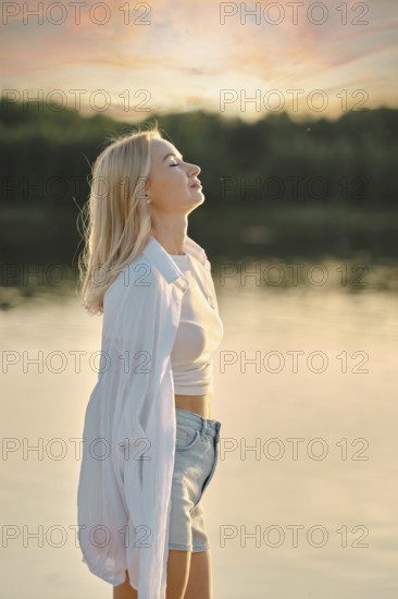 A woman stands by the calm lake, feeling the gentle breeze as she admires the colorful sunset sky. She wears a light shirt and shorts, embracing the peaceful atmosphere of the evening
