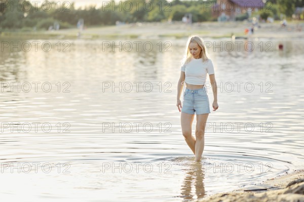 A young woman in a white crop top and denim shorts steps delicately into the clear water of a beach