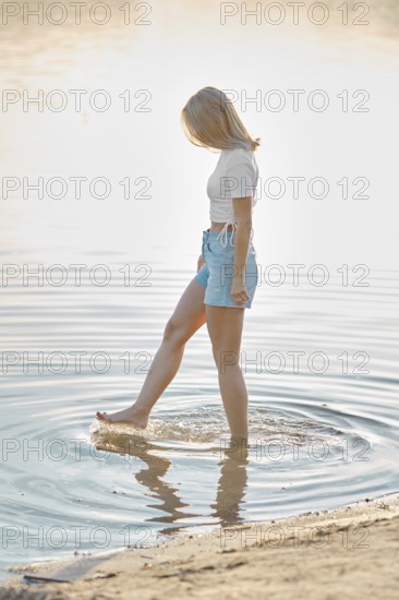 A woman stands in shallow water at a beach during golden hour, playfully splashing around with her feet. The soft light creates a serene atmosphere while she enjoys the tranquility of nature