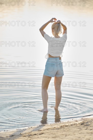 A woman stands in shallow water, playfully wringing her hair as sunlight glimmers on the surface. The serene environment showcases natural beauty with ripples forming around her feet