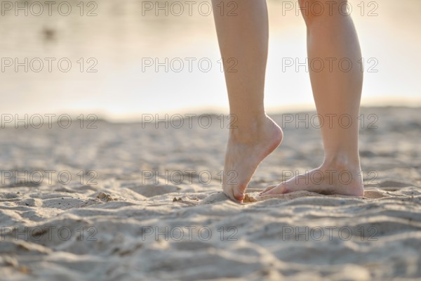 A person stands on soft, golden sand with bare feet, enjoying the warm texture as the sun sets in the background. The peaceful beach atmosphere enhances the sense of relaxation and freedom