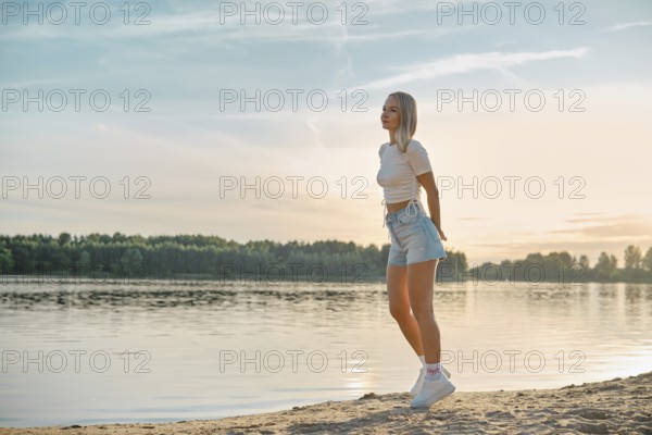 A young woman walks leisurely along the sandy lakeshore as the sun sets behind the trees. She wears casual summer attire, embodying the peaceful atmosphere of the evening