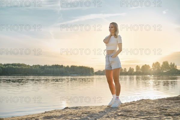 A young woman relaxing on the sandy beach near a calm river at sunset. She wears a casual outfit with shorts and a crop top, enjoying the serene view of the evening sky