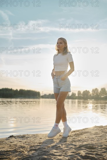 A young woman walks on tiptoes on a sandy shore as the sun sets over the lake. Her casual outfit complements the serene atmosphere, with soft light illuminating the scene