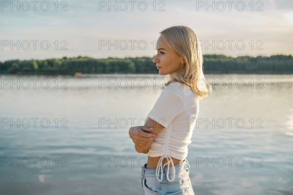 A blonde woman stands in profile by a calm lake, gazing thoughtfully into the distance. The sun sets, casting warm hues over the water as the serene landscape reflects a peaceful atmosphere