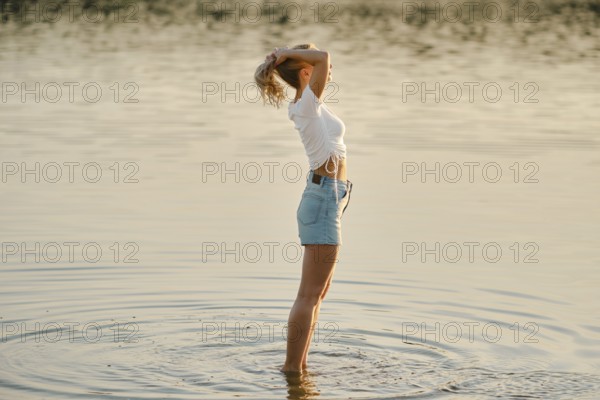 A woman stands in shallow water in profile, basking in the warm glow of sunset. She raises her hands and gathers her hair in a ponytail