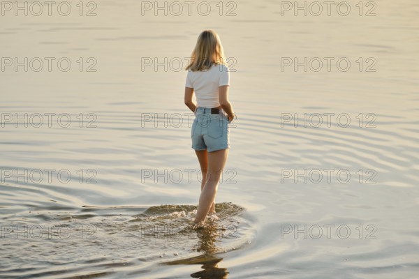 A woman with blonde hair wades through calm, shallow water as the sun sets behind her, creating a warm glow. The peaceful atmosphere invites relaxation and reflection in nature