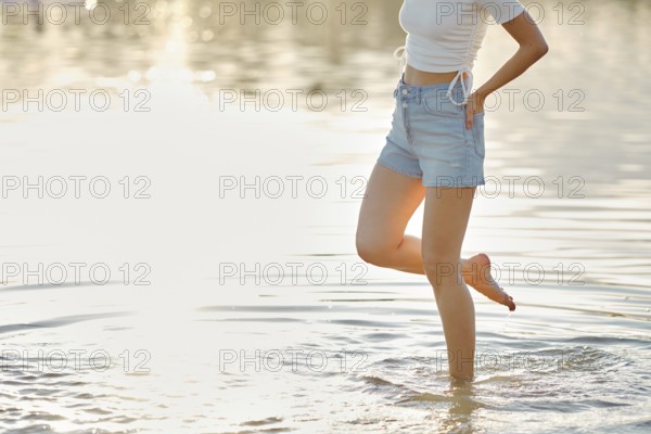 Unrecognizable woman steps carefully in the clear water, enjoying the warmth of the sun on her skin. Wearing a light top and denim shorts, she experiences the refreshing sensation of the water