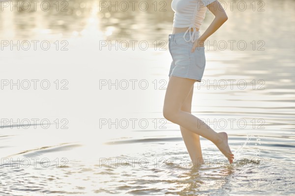 Unrecognizable woman stands in shallow water, playfully splashing while enjoying a serene sunset at a lake. The soft sunlight reflects off the water, creating a tranquil atmosphere around her