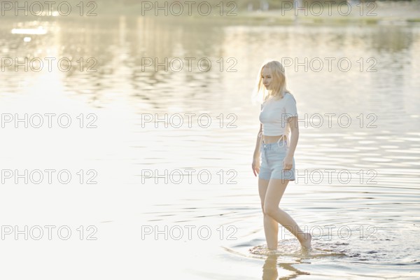 A young woman with light blonde hair walks barefoot in shallow water at sunset. The serene lake reflects the golden light, creating a calm atmosphere as she enjoys the peaceful surroundings