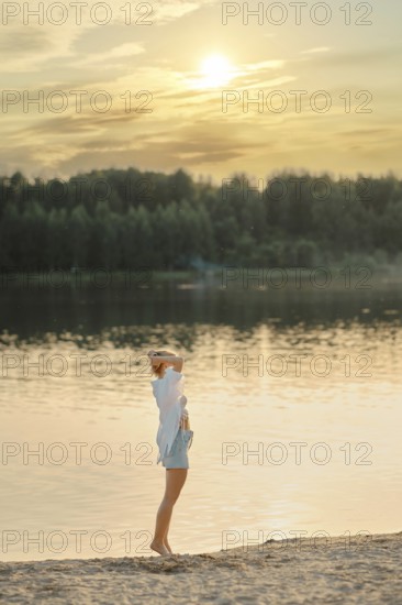 A woman stands at the edge of a tranquil lake during sunset, gazing across the water. Warm golden light reflects off the surface while trees create a peaceful backdrop