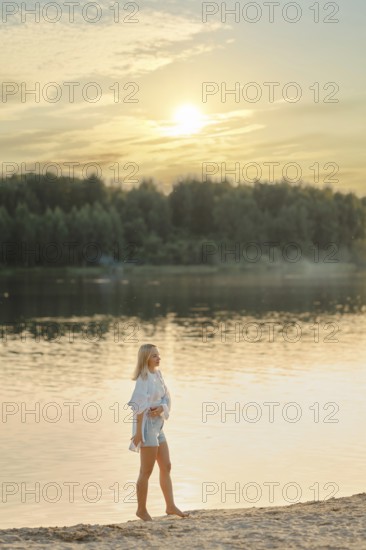 A woman strolls barefoot along the sandy shore of a serene lake during sunset. The sun sets behind trees, casting a warm glow over the water and creating a peaceful atmosphere
