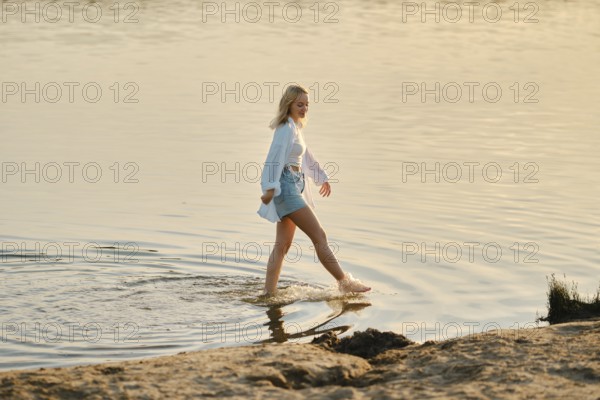 A young woman with blonde hair strolls through shallow water during sunset, her feet gently making ripples