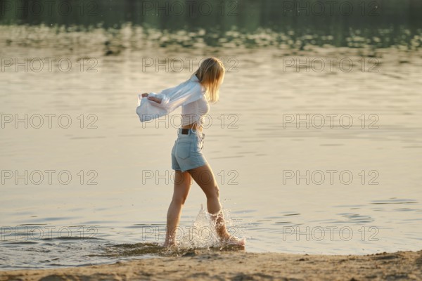 A young woman strolls through shallow water at a peaceful lakeshore creating splashes with her feet during sunset