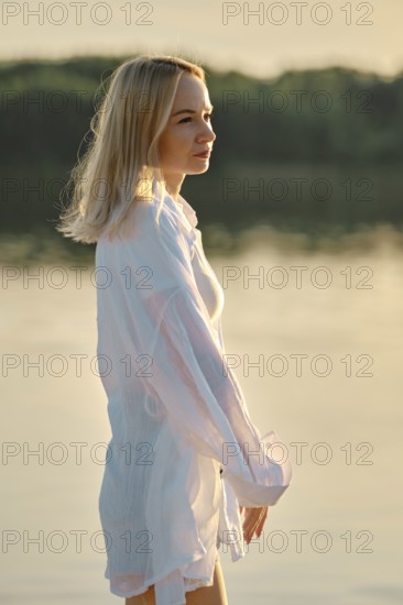 Woman with light blonde hair gazes thoughtfully into the distance while standing near tranquil water. The sun sets behind trees, creating a warm, golden light that enhances the peaceful atmosphere