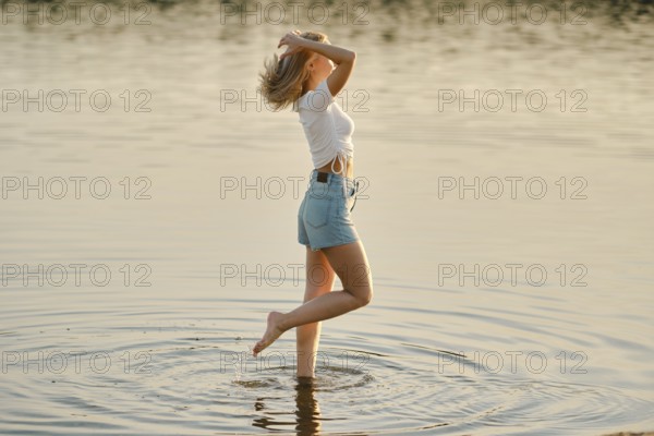 A young woman is joyfully splashing in the shallow waters of a serene lake. The sun is setting, casting a warm glow over the scene. She lifts her hair while playfully moving through the water