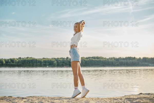 A young woman walking along a tranquil lake at sunset, dressed in casual summer attire. She stretches her arms blissfully, surrounded by lush greenery and calm water reflecting the sky