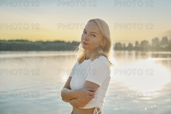 Cute woman stands thoughtfully by the lake, gazing into the distance as the sun sets behind her. The water reflects the warm colors of dusk, creating a peaceful atmosphere for relaxation