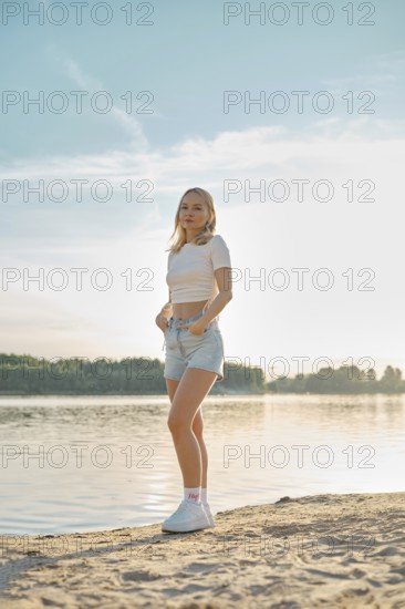 A young woman stands on a sandy beach, facing the camera. She wears a casual outfit of a cropped top and denim shorts, with the sun setting beautifully behind her, creating a warm glow