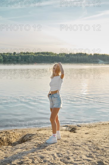 A young woman stands on the sandy shore of a calm lake, admiring the sunset. Her relaxed pose and casual outfit create a tranquil atmosphere, highlighting the beauty of nature around her