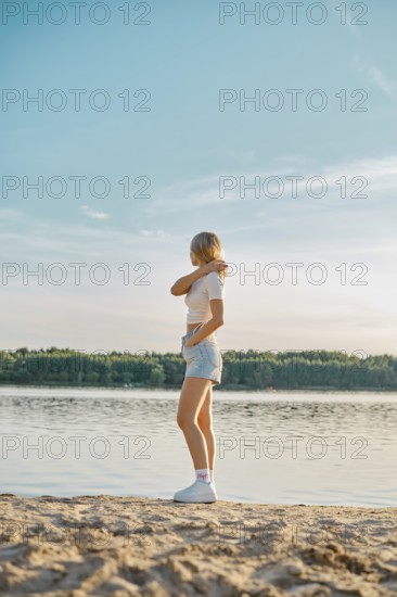 A young woman stands on the sandy shore of a tranquil lake, gazing at the water while wearing casual summer attire. The sun sets in the background, painting the sky with warm colors