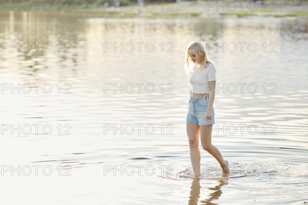 A young woman with blonde hair strolls through shallow water in a lake during sunset. The bright colors reflect on the surface, creating a calm atmosphere as nature surrounds her