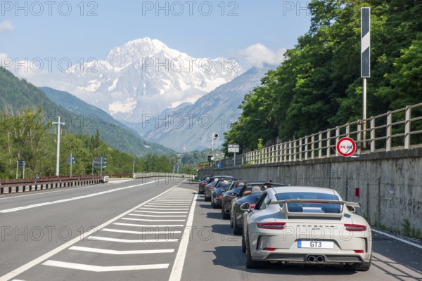 View of the highest mountain in the Alps Mont Blanc in the foreground several Porsche sports cars in a row on Strada Statale 26 SS26, La Salle, Autonomous Region of Valle d'Aosta, Italy