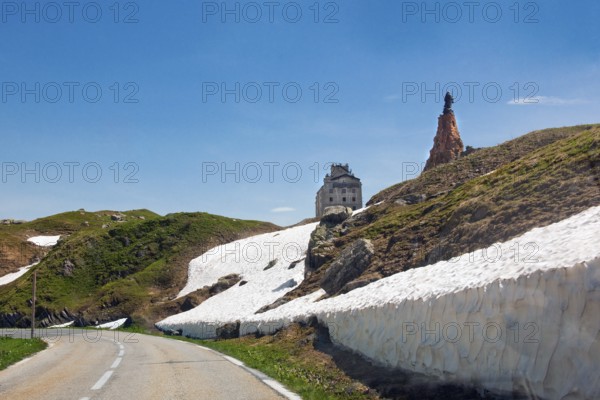 View from last bend of alpine road pass road mountain road in front of pass summit of Little Saint Bernard Pass Colle del Piccolo San Bernardo, Col du Petit Saint Bernard Little Saint Bernard Pass in Graian Alps, top right statue of monk Saint Bernard of Menthon on large pedestal made of natural stones, top centre historic renovated reopened hotel Hospice l'Hospice de Petit Saint Bernard, Département Savoie, France