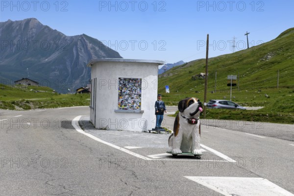 Disused former border hut for passport control at the border crossing between France and Italy, in the foreground figure of a St Bernard dog with a barrel around its neck, behind it figure of a French border official, Graian Alps, Département Savoie, France, Valle d'Aosta, Italy