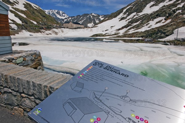 Tourist information board of Great St. Bernard Pass, behind it pass summit with icy lake Lago di Gran San Bernardo, Valais, Switzerland, Valle d'Aosta, Italy