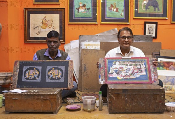 Miniature painters, aged 50 and 52, show paintings of elephant and royal city palace in studio, Udaipur, Rajasthan, India