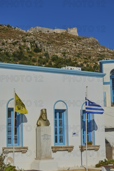 Bust in front of a white building with blue windows, Greek flag and yellow flag in the foreground, hill in the background, Agia Marina, Leros, Dodecanese, Greek Islands, Greece
