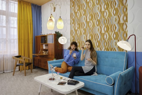 Two friends are seated on a vibrant blue sofa in a stylish living room filled with natural light and applying make-up