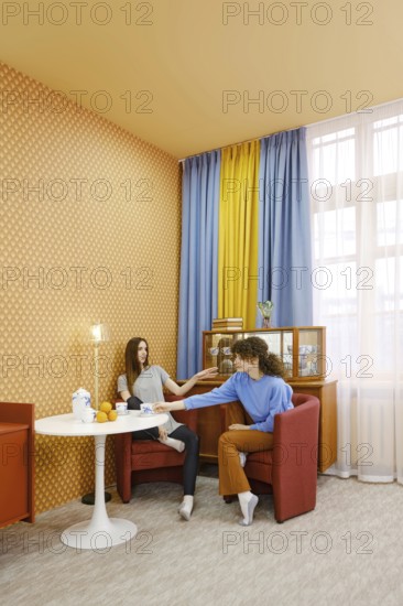 Two women are seated in a bright room, engaged in a lively discussion. One woman reaches out with a playful expression while the other listens attentively