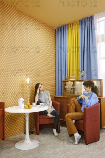 Two friends are sitting comfortably in a living room with bold wallpaper and bright curtains. They engage in a conversation, discussing female problems