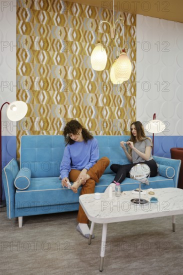 Friends spend a pleasant afternoon in a modern lounge, sitting on a turquoise sofa. They engage in conversation while one polishes her nails and the other removes dead skin of heel