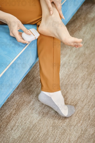Unrecognizable woman use electric gadget to peel her heel from rough or dead skin while sitting on a blue couch in a cozy living room
