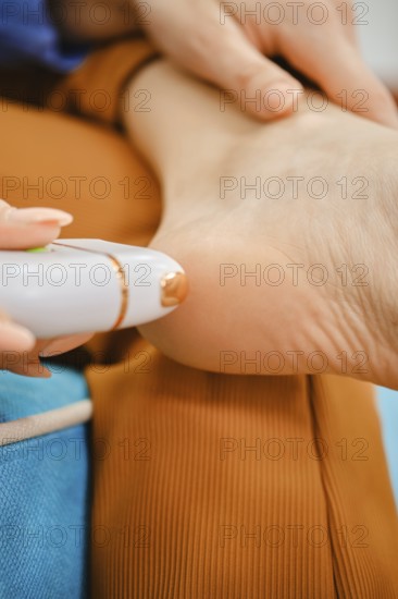 A woman is treating her foot with a device designed for removing rough skin. She sits comfortably with her foot rested, focusing on achieving smooth and soft soles at home, enjoying personal care