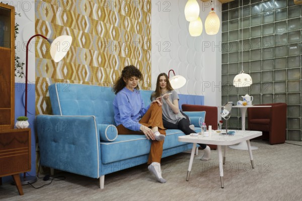 Two women are seated on a blue sofa in a living room, enjoying each other's company while engaging in beauty procedures