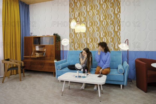 Two women sit on a turquoise sofa in a stylish living room and removing makeup. The vibrant decor adds warmth to their engaging conversation about an important topic