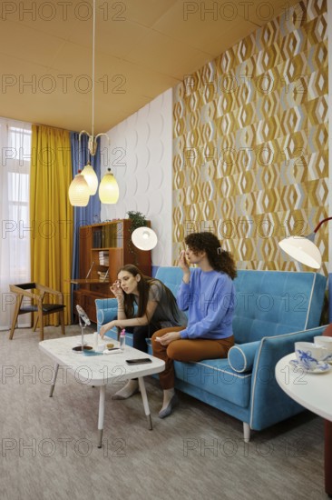 Two women sit comfortably on a bright blue sofa in a contemporary living room. They are engaged in a light conversation while applying make-up and enjoying each other's company