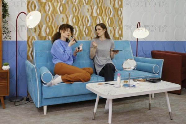 Two friends are sitting on a blue sofa and discussing makeup. The room features stylish decor, including a patterned wall, lamps, and a coffee table