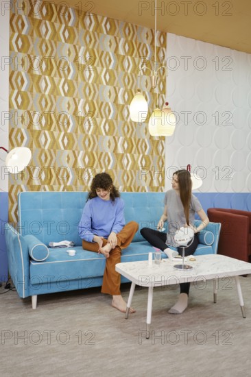 Two young women are engaged in skin care procedures at home sitting on a cozy sofa. One woman rubs the cream in the feet