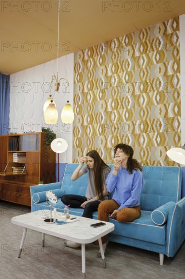Two women remove makeup from their faces sitting at home on a blue couch in a cozy living room, sharing a light conversation