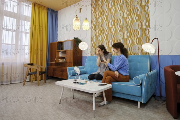In a bright living room adorned with curtains, two friends sit on a blue sofa, chatting and laughing. One woman applying collagen patches on face skin under her eyes